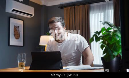 Young man in white t-shirt smiling and engaging in a video call on tablet, sitting at cozy home desk with papers and tea. Stock Photo