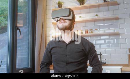 Bearded man in black shirt uses a VR headset in a bright modern kitchen, enjoying immersive digital content or virtual exploration. Stock Photo