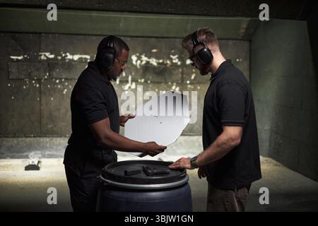 Two young men, Black and Caucasian, standing in indoor shooting range examining paper target with bullet holes while wearing protective ear muffs and black shirts Stock Photo