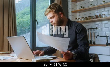 Focused bearded man sits at desk by window, reviewing printed documents while using a laptop in a cozy indoor workspace. Stock Photo