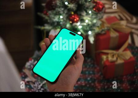 hand holding a smartphone with a green screen display against a Christmas tree and red gifts background Stock Photo