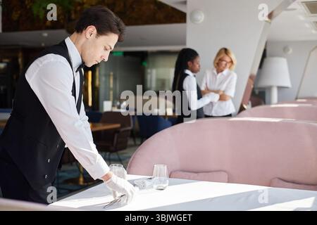 Young Caucasian Man Setting Table While Two Female Coworkers Discussing Work Stock Photo