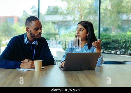 Two colleagues are engaged in a discussion about accounting strategies while seated at a desk in a modern office. With a laptop open in front of them, Stock Photo