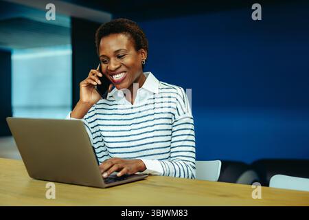 A smiling African businesswoman is engaging in a phone conversation while working on her laptop in an office setting. Her joyful expression captures a Stock Photo