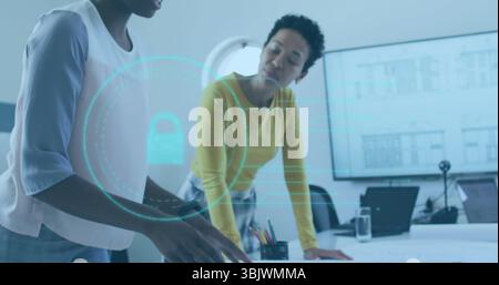 Leaning two businesswomen examining documents at office table, with laptop and holographic padlock Stock Photo