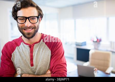 Bearded man wearing black glasses and red-and-gray henley standing in bright office near laptop Stock Photo