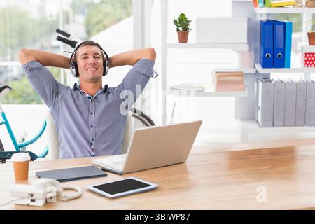Male office worker wearing headset leaning back in office using laptop and holding coffee mug Stock Photo