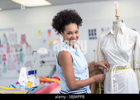 African American woman fitting dress on mannequin in studio with sewing machine and tape measure Stock Photo