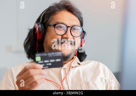 Mid adult smiling Asian businessman in office with headphone and using credit card to pay online. Selective focus. Stock Photo