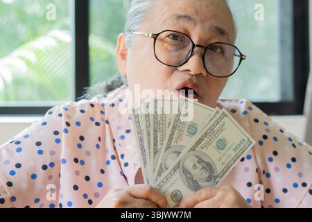Senior grey-haired Asian woman holding 100 US Dollar banknotes with smiling friendly gesturing excellent face. Close-up. Senior wealth concept. Stock Photo