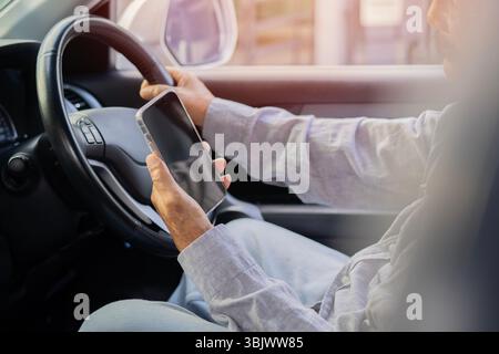 Side view of old man drives his car while using or looking on mobile phone. Dangerous driving on the road Stock Photo