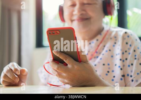 Modern Asian elderly woman laughing watching a video on the internet with her smartphone and headphones at home. Selective focus. Stock Photo