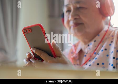 Side view of an elderly Asian woman wearing headphones and smiling while holding a cell phone. Retirement lifestyle concept. Stock Photo