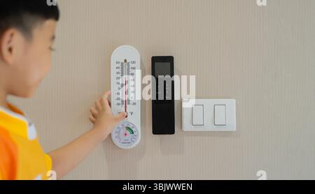 Boy looking at a thermometer, nearby is a black air conditioner remote and light switch on the wall background. Stock Photo