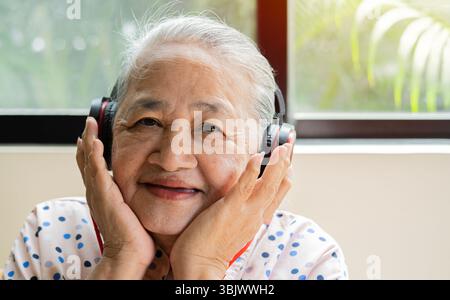 Portrait of mature Asian woman smiling and looking to the camera while listening to music on headphones. Stock Photo