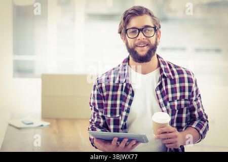 Bearded man wearing checkered shirt holding tablet and coffee cup near stacked boxes in bright room Stock Photo