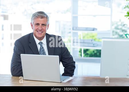 Silver laptop sitting on wooden desk near large window, white shelving holding folders and plant Stock Photo