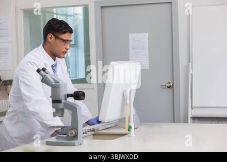 Male researcher wearing lab coat, gloves typing on PC at lab bench with microscope, pipette stand Stock Photo