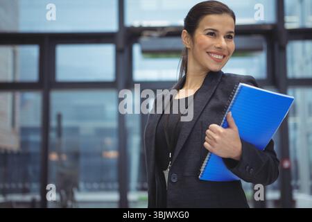 Businesswoman standing in lobby holding blue notebook and wearing lanyard badge smiling, copy space Stock Photo