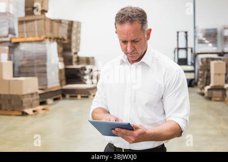 Mature adult man standing in warehouse checking tablet near forklift and cardboard boxes on pallets Stock Photo