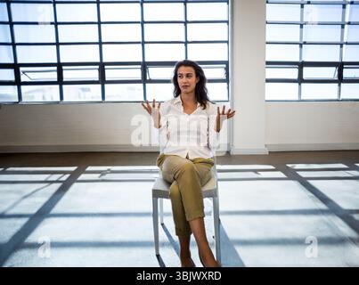 Woman in mid-thirties sitting on white chair in loft, casting grid window shadows across floor Stock Photo