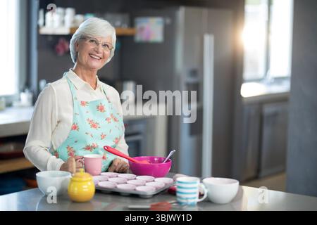 Senior woman pouring cupcake batter into muffin tin on countertop wearing floral apron, copy space Stock Photo