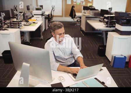 Middle-aged man in business casual attire typing on laptop at desk in open-plan office with monitor Stock Photo