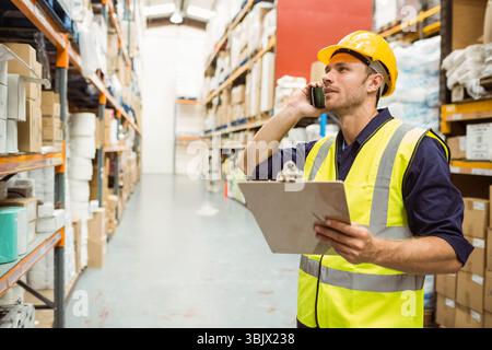 Man wearing vest and helmet checking shelves in warehouse aisle with clipboard, phone, copy space Stock Photo