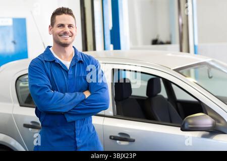 Male technician in blue coverall smiling while standing beside silver hatchback in repair shop bay Stock Photo