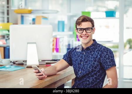 Man in his twenties holding smartphone while typing on keyboard at desk with monitor, copy space Stock Photo
