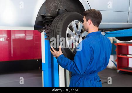 Mechanic positioning removed car tire beneath lifted silver vehicle on hydraulic lift in shop Stock Photo