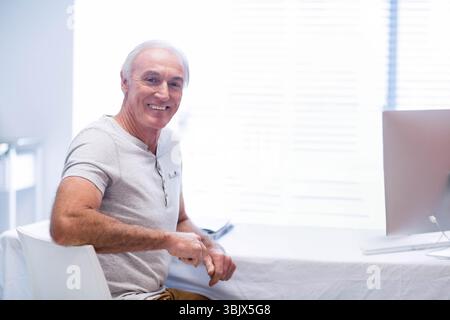 Smiling senior man working on desktop computer in home office with smartphone and window blinds Stock Photo