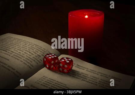 A dark, atmospheric composition featuring two red translucent dice placed on an open book illuminated by the warm glow of a red candle. The soft light Stock Photo