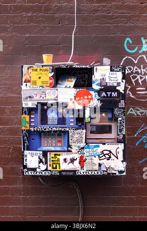A vintage ATM covered in graffiti and stickers installed on a brick wall with urban street art and colorful designs surrounding it Stock Photo
