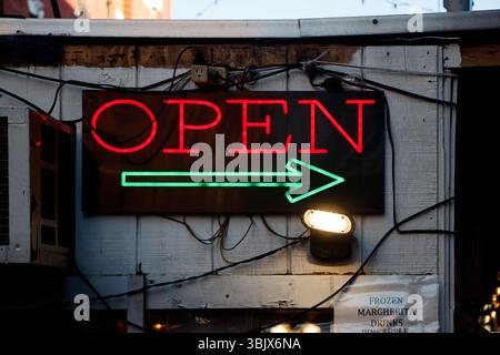 Neon sign displaying the word 'OPEN' with a green arrow against a wooden wall backdrop, illuminated at night Stock Photo
