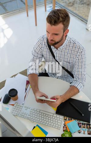 Bearded man checking smartphone at round desk near glass windows with computer, tablet, coffee cup Stock Photo