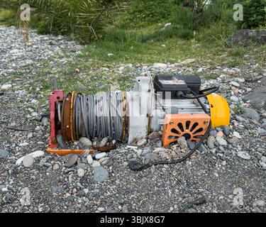 Abandoned electric winch with steel cable lying on rocky shore, corroded and unused, symbol of coastal decay. No people. Stock Photo