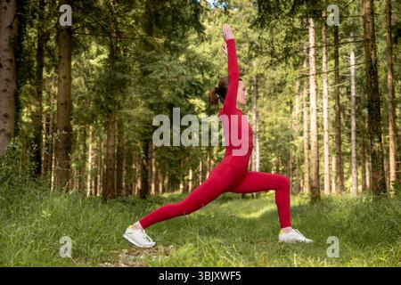 Fit Sporty Woman Practicing Outdoor Yoga in Forest Performing Warrior Pose in Red Outfit Stock Photo