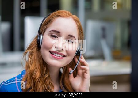 Red-haired support rep using headset and blue plaid shirt smiling at open-plan office environment Stock Photo