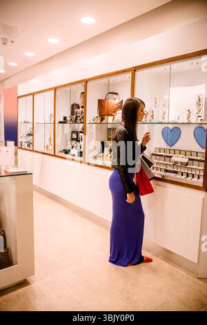 Female shopper wearing leather jacket near glass jewelry display, holding red and striped bags Stock Photo