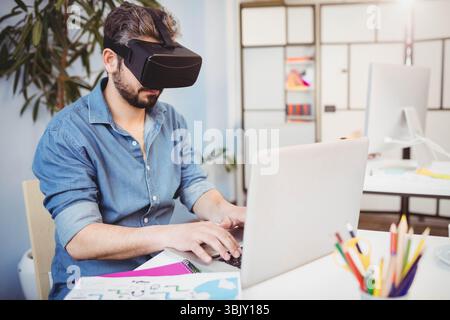 Man wearing black VR headset typing on laptop with sketch papers and colored pencils at desk Stock Photo