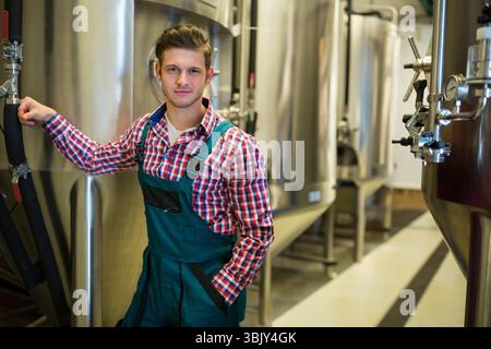 Male brewery worker in overalls standing near fermentation tanks at brewery with gauges, copy space Stock Photo