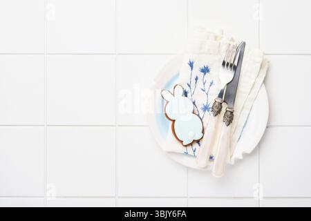 Easter table setting with gingerbread rabbit, napkin, knife, fork and plate on white ceramic tile background. Elegance dinner. M Stock Photo