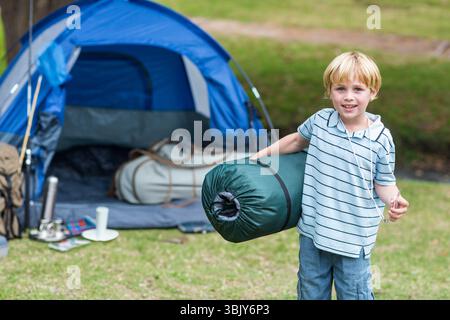 Boy carrying rolled sleeping bag while standing on grassy campsite by blue dome tent, copy space Stock Photo