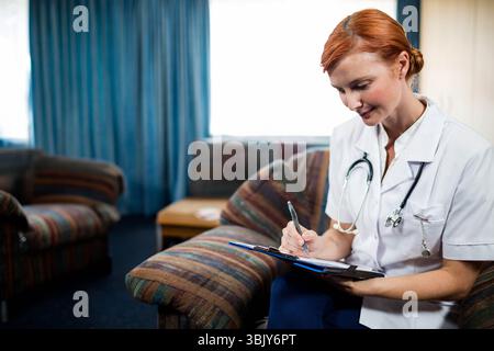 Red-haired female health worker writing on clipboard in lounge wearing white uniform, copy space Stock Photo