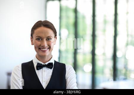 Female server smiling and standing in restaurant lobby wearing white shirt black vest bow tie Stock Photo
