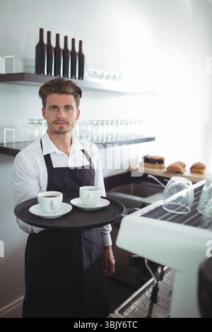 Male barista in white shirt, black apron holding tray with two coffee cups on coffee counter Stock Photo