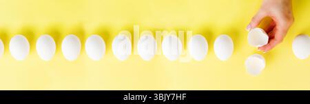 White raw chicken eggs lying in row on a yellow trendy background and female hand holds broken egg. Easter minimal creative comp Stock Photo