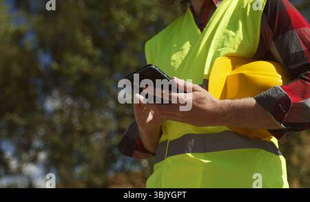 Builder with hard hat and high vis jacket using smartphone. Close-up. Stock Photo