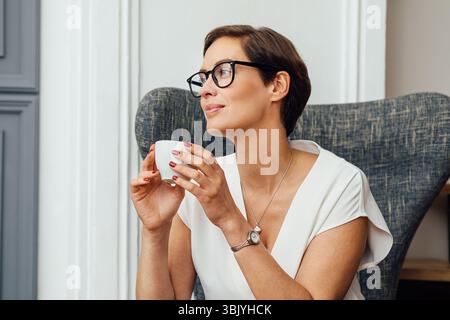 Businesswoman sitting on armchair in her apartment and holding a cup of coffee Stock Photo